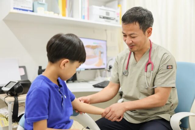 医師が男の子の腕に手を添えて話しかける診察室の様子